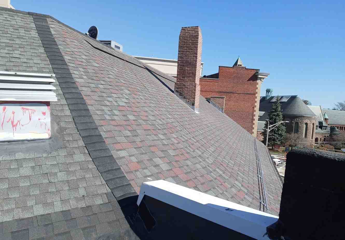 Steep rooftop with gray shingles, a brick chimney, and a painted vent. Nearby, red-brick buildings and a few trees under a clear blue sky.